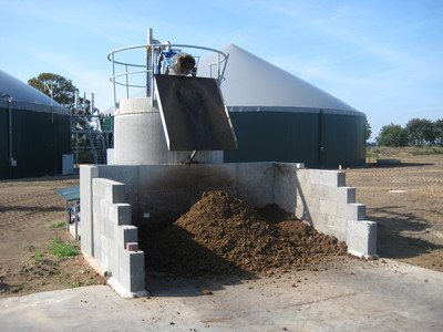 Das Bild zeigt einen Separator, der auf einem Podest steht. Darunter befinden sich separierte Gärprodukte auf eine Lagerplatte. Im Hintergrund ist eine Biogasanlage zu sehen.